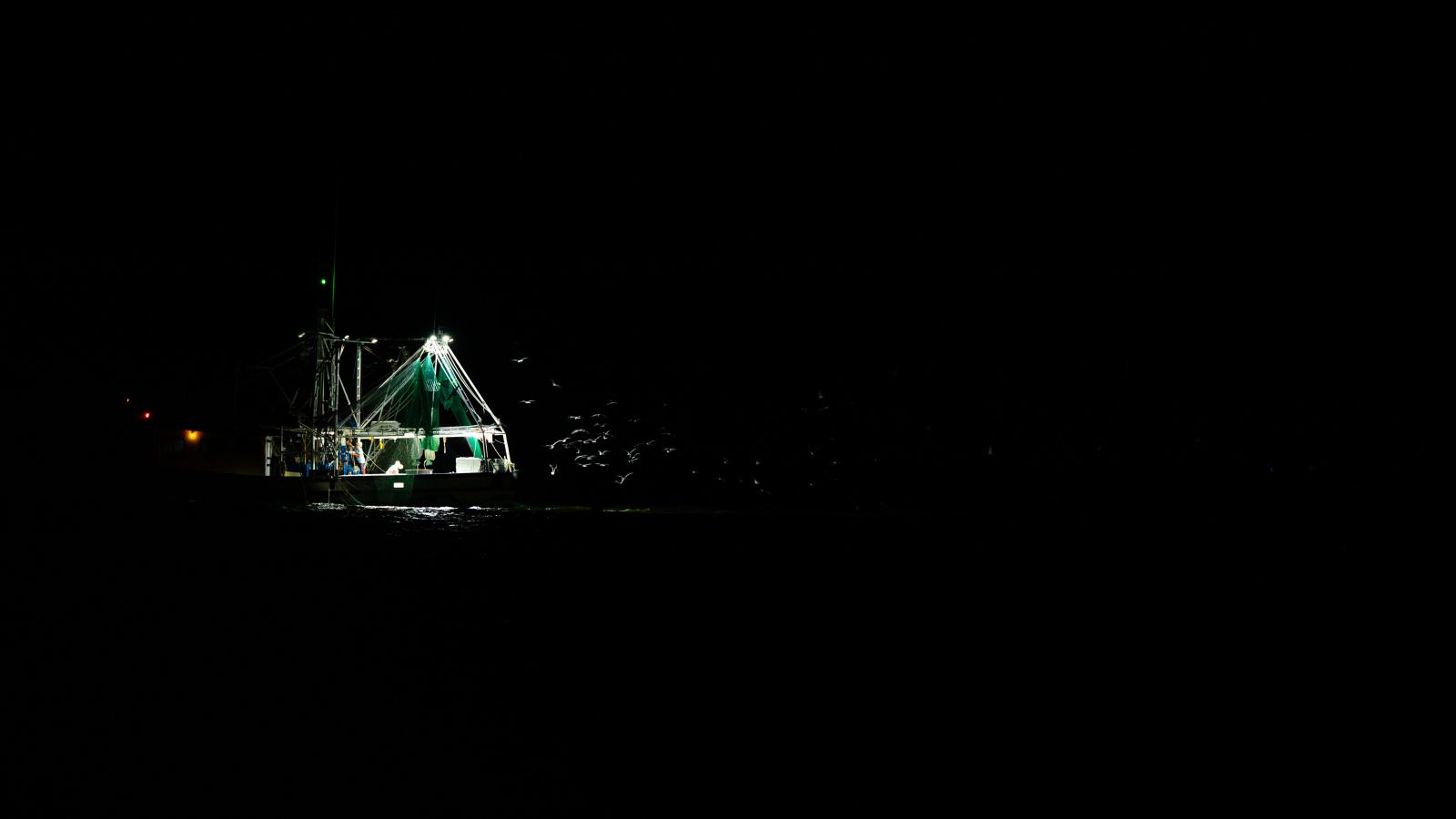 A wide shot of a fishing boat in the middle of the night surrounded by darkness.